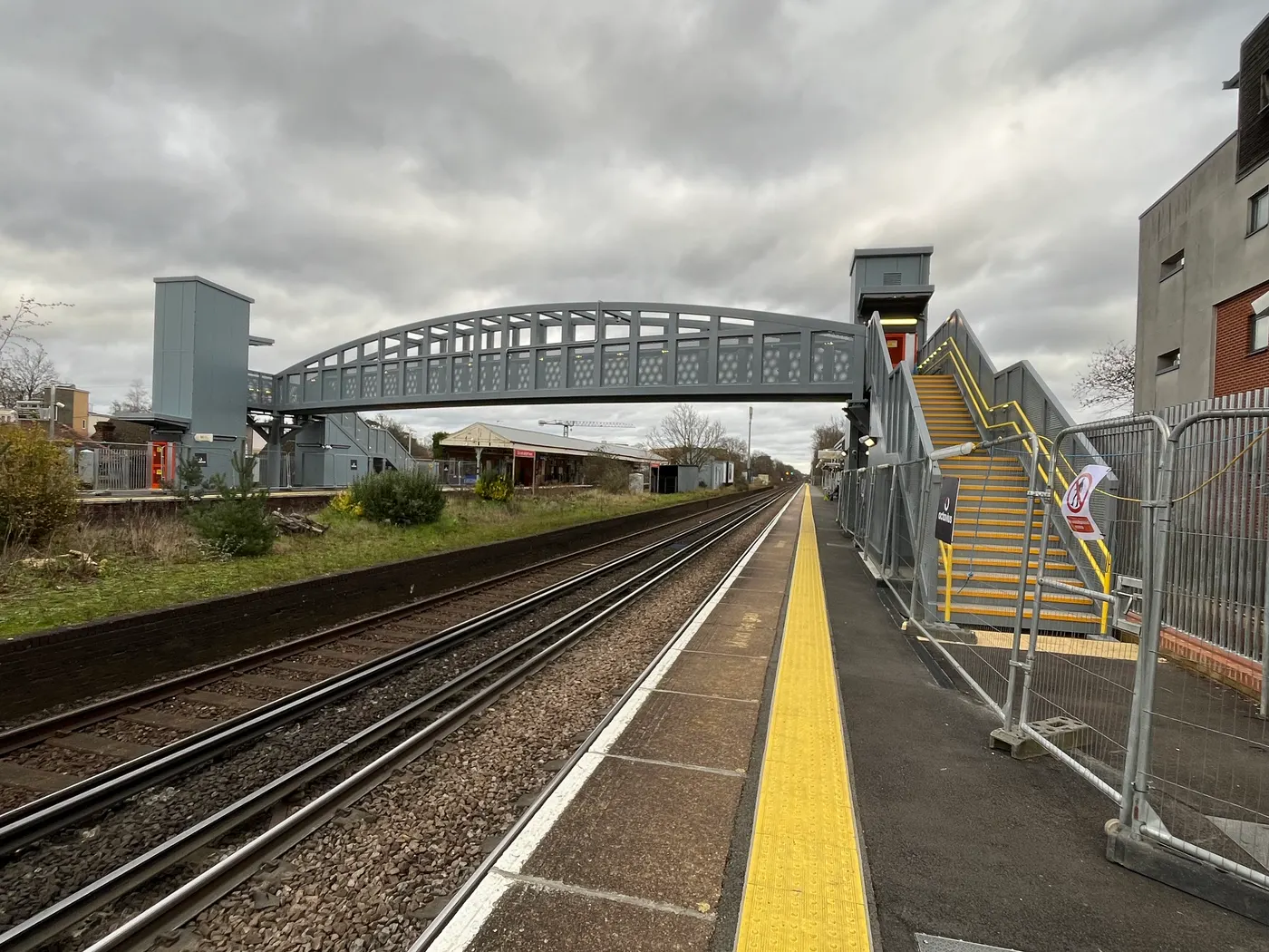 The new arched steel footbridge at Walton-on-Thames station, with a lift shaft and a new staircase with yellow handrails still enclosed by contractors' fencing