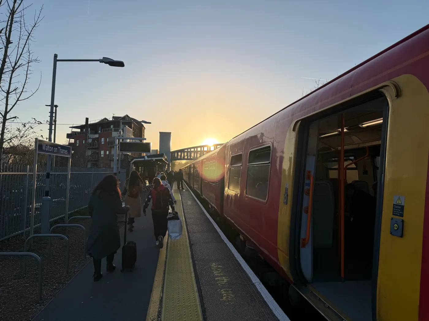 A red South Western Railway train standing at Platform 2 at Walton-on-Thames with its doors open at sunset, as passengers walk away along the platform towards the footbridge